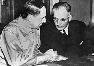 Two men seated at a table side by side talking. One is wearing a suit, the other a military uniform.