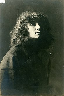 A young white woman with loose curly hair, wearing a plain dark garment, photographed mainly in shadow.