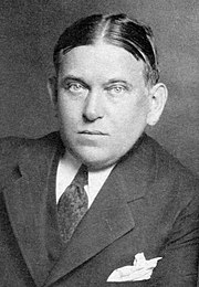A photographic portrait of critic H. L. Mencken. His hair is parted in the middle, and he appears to be leaning on his left arm. He is wearing a dark tie and a dark suit with peak lapels. A white handkerchief is visible in his suit pocket.