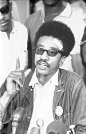 Un jeune homme afro-américain.  Il parle dans un microphone et fait des gestes;  il porte des lunettes de soleil.