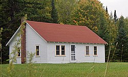 A gabled, one-story wood-frame cabin with forest rising immediately behind