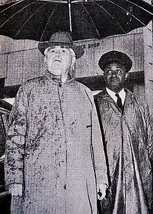 John Lewis en pardessus et chapeau sous parapluie tenu par James Lewis Jr. en uniforme de chauffeur.