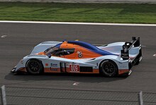 Photographie d'une voiture de sport-prototype, orange, bleu ciel et bleu foncée, de haut et de profil, sur une piste.