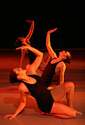 Three women in black leotards, each kneeling on one leg with the other foot on the floor, leaning back to support herself on one hand, and looking at the other hand, which is raised