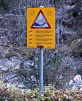 Panneau jaune avec avertissement en plusieurs langues. Il dit : « Danger… Il est dangereux de stationner dans le lit de la rivière, l'eau pouvant monter brusquement et à tout moment par suite du fonctionnement des installations hydro-électriques. … Aussi par beau temps. »