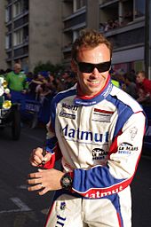 A smiling man in his late 20s wearing black rectangular sunglasses and white, blue and red racing overalls. He is holding a ballpoint pen in his right hand and there are a crowd of people behind him up against blue adveristing metal fences.