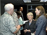 Two adult women talk with an older white-haired man in camouflage inside a dark room.