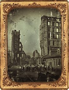 A crowd of people gather in front of the ruins of the Lindell Hotel after the fire on 30 March 1867. Two sections of the hotel rise above a pile of rubble. Two horses can be seen in the bottom-right corner. The building next to the hotel is labeled with two signs that read "Globe Exchange" and "Beer Saloon, Cigars". Ruins of the Lindell Hotel, after fire of 30 March 1867.jpg
