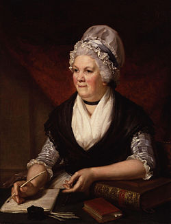 Half-length portrait of an elderly woman sitting at a desk surrounded by books and papers and holding a quill