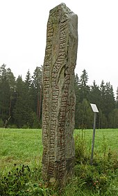A vertical inscribed stone rising from gras with treest in the background.