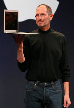Crotch-high portrait of man in his fifties wearing blue jeans and a black turtleneck shirt, carrying an open laptop computer in his right hand, large Apple logo cut off behind him
