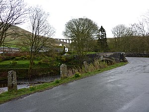 In the foreground, a narrow road going over a stone bridge with trees without leaves at the sides. In the background, steep hills with a railway viaduct straddling a gap