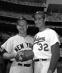 "Two men pose for an image; the shorter one is dressed in pinstripes and a wears a ballcap with an interlocking 'NY' while the taller one wears a plain uniform with 'Dodgers' across the front and a ballcap with an interlocking 'LA'."