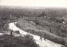 Schwarzweiss-Foto einer Gruppe von Soldaten, die auf einem Weg in einem spärlichen Dschungel marschieren.