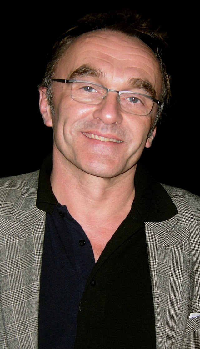 The photograph of a balding brunette man who is smiling. He wears a black shirt and a beige blazer.