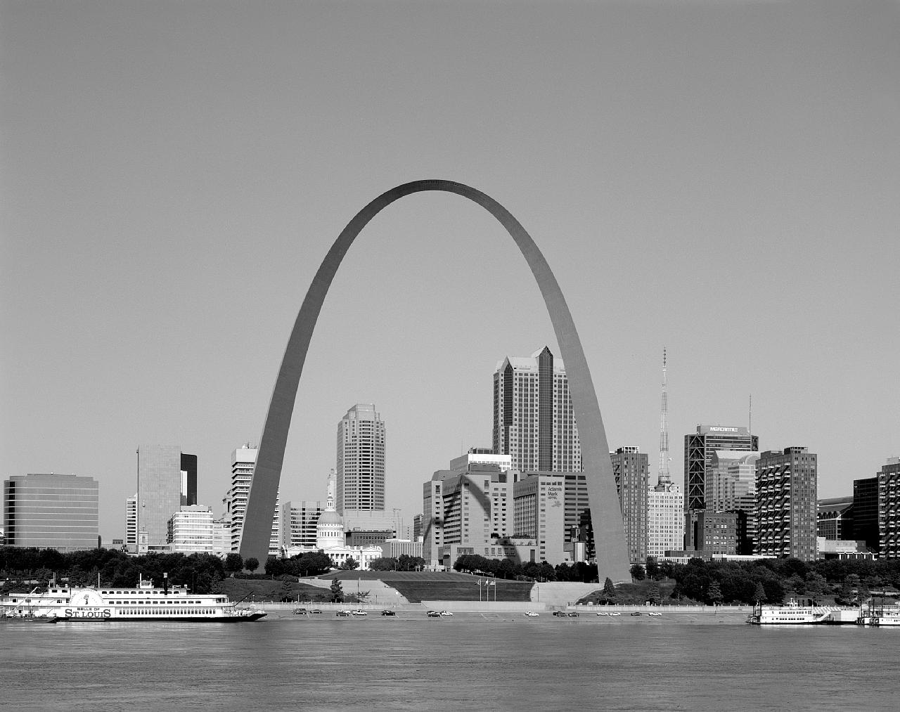 Photographie en noir et blanc d’une arche monumentale avec un fleuve au premier plan et des immeubles à l’arrière-plan.