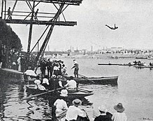 Photographie noir et blanc : des barques sur l'eau et un homme plongeant d'une structure en bois.