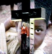 A faraway image of a blond woman in front of a giant, mirrored cross. She is wearing a red shirt and brown pants. On her head a crown made of thorns is visible. Behind the cross, the face of an African child is visible on a ginat screen