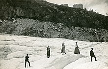 Photo en noir et blanc de touristes sur un glacier et, sur la crête en arrière-plan, trois bâtiments de taille inégale.
