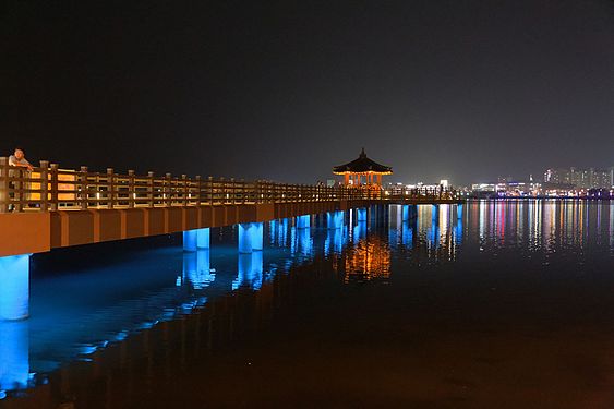 Ein Pavillon, der über einen Steg vom Ufer aus begehbar ist, im Brackwassersee Cheongchoho. Im Hintergrund ein Teil der Stadt.