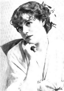 A young white woman with dark eyes and curly hair in an updo, seated, chin resting on one hand