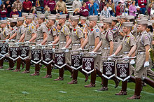 Row of students in uniform with knee-high boots.  Each holds a bugle that dangles a pennant.