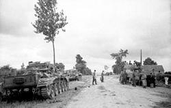 Two tanks are parked on a grass verge to the left of a road while another tank is parked on the right side being inspected by a group of men. Two men are standing in the middle of the road.