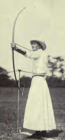 A white woman archer wearing a white hat, blouse, and ankle-length skirt