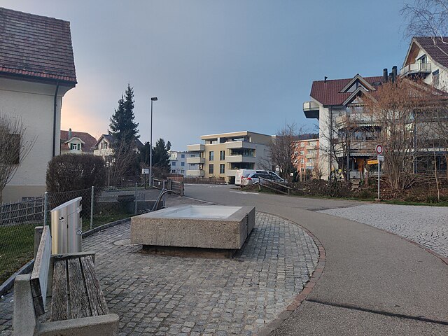 The concrete fountain which today is located at the intersection of Zihlstrasse and Gstaldenstrasse in Hinwil was designed by Max Vogt as part of the Hinwil railway station. It has since been moved away from the railway station and placed here. The photo shows the fountain on the western side of the road, looking north into Gstaldenstrasse. The fountain is empty, presumably because water supply is turned off during winter