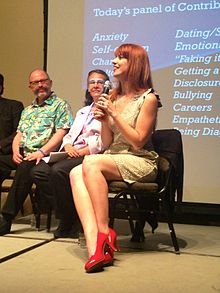 A woman with long, straight ginger hair sits facing left. She wears a white sleeveless dress and red high heels and is holding a microphone. Two older-looking people, a man and a woman, are visible in the background.