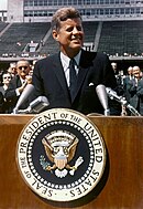 President Kennedy is pictured speaking behind a podium. Rice University's stadium is visible behind him.