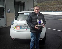 Homme de face tenant un trophée composé entre autres d'une sphère bleue et du sigle FIA sur son coté gauche.