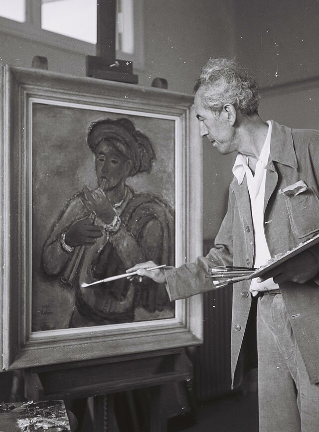 תצלום שחור-לבן. ראובן רובין מצייר בסטודיו שלו. רובין ניצב מימין לתמונה שהוא מצייר, הניצבת על כן ציור בשמאל התמונה.