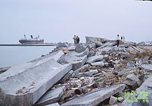 Slightly elevated photo showing a pile of rubble near the coast, with a few onlookers standing next to it.