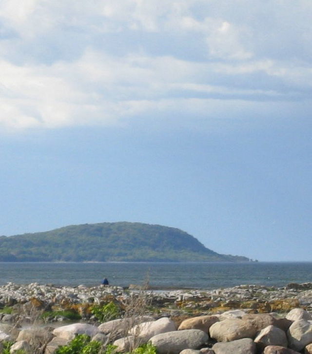 A grey-blue body of water separating smooth, large, light-coloured rocks in the foreground and a hazy green hill in the background, all under a blue sky with white clouds.