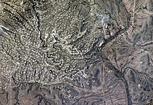 Une photographie regardant vers le bas sur un terrain vallonné et sans arbres avec une ville en haut à droite et la fine ligne d'une rivière traversant de haut en bas sur la droite