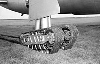 The experimental tracked landing gear unit of an XB-36, consisting of 2 flexible tracks supported by wheels, is pictured stopped in short grass with ruts trailing behind it.