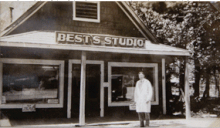 Na černobílé fotografii je muž oblečený jako malíř, stojící před dřevěným domem, na kterém je cedule s nápisem BEST´S STUDIO.