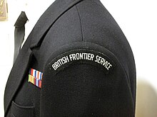 Side view of the shoulder of a dark-coloured uniform with the words "British Frontier Service" visible on a shoulder patch and a row of medal ribbons visible on the front left breast above a pocket. British frontier service uniform.jpg