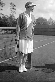 Nainen, joka katsoo poispäin kamerasta, tennismaila oikeassa kädessään ja värillinen pusero ja kaikki valkoiset vaatteet, jotka tämä kuva on mustavalkoinen