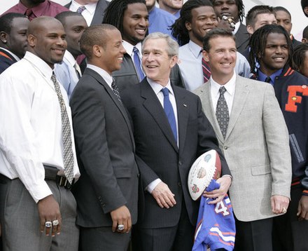 Photo depicts a smiling Chris Leak, quarterback of the Florida Gators, together with his Florida Gators football teammates, coach Urban Meyer, and U.S. President George W. Bush, who is holding a Gators football jersey and commemorative football.