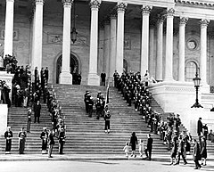 Body bearers carrying the casket of President Kennedy up the center steps of the United States Capitol Building, followed by a color guard holding the flag of the President of the United States, and the late President's widow, Jacqueline Kennedy and her children, Caroline Kennedy and John F. Kennedy Jr., on November 24, 1963.