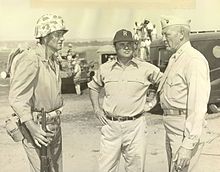 Actor John Wayne in military uniform with helmet and gun stands by a man in a cap and a man in uniform.