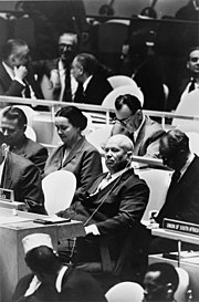 An older man sits among United Nations delegation tables, looking at the camera.