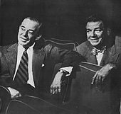 Двое мужчин в деловых костюмах сидят в театральных креслах и дружелюбно обсуждают то, что смотрят.