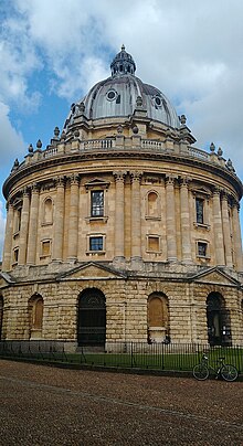 Photo of the Radcliffe Camera from street level showing the Rotunda shaped library with grey top and cream bricks. The railings around the outside have bicycles against them.