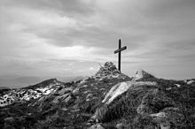 Schwarz-Weiß-Fotografie eines hölzernen Gipfelkreuzes und eines Steinhaufens mit bewölktem Himmel.