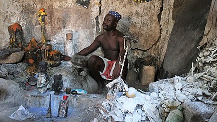 Vodou-pappi tarpeistoineen, kuten figuriineja ja fetissejä. Keskellä edessä asen-alttari.