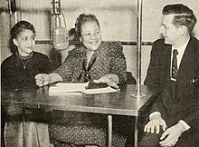A photograph of two women and a man seated at a table with a hanging microphone in the center and papers on the table