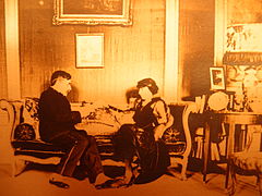 Zengin bir şekilde döşenmiş oturma odasında kanepede oturan bir kadın ve bir adam.  Tozluklu adam bir el yazmasını okuyor.